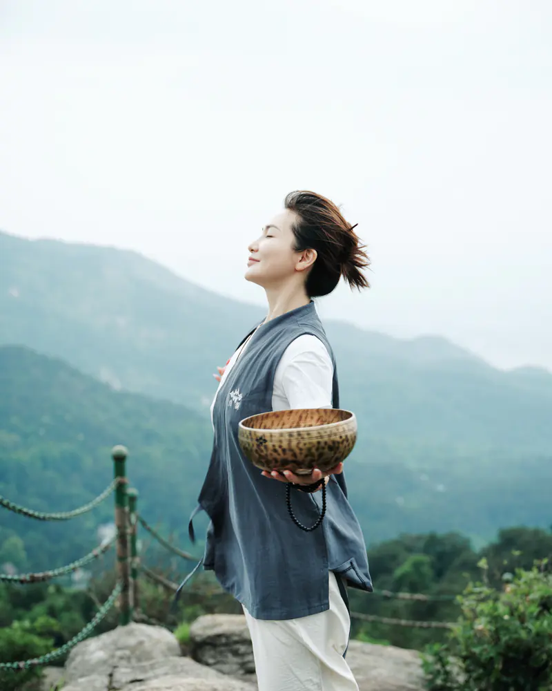 A person naturally holds a metal singing bowl in a mountain setting, placing the instrument in an Eastern living context